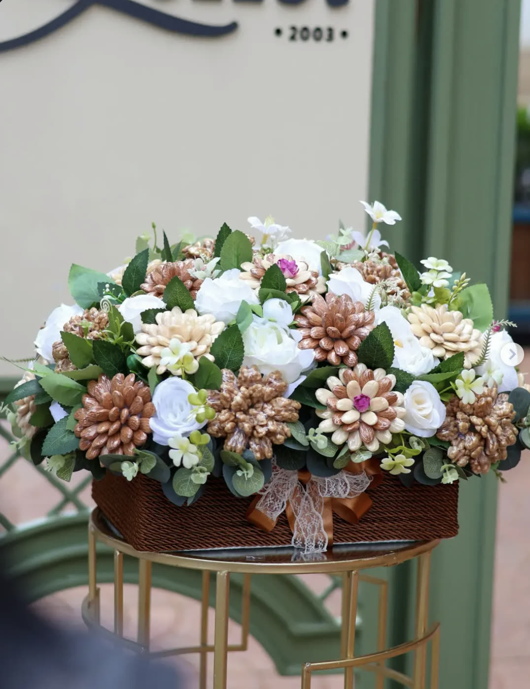 Wicker basket of white roses and gold pine‑cone flowers on a stand outside the gallery