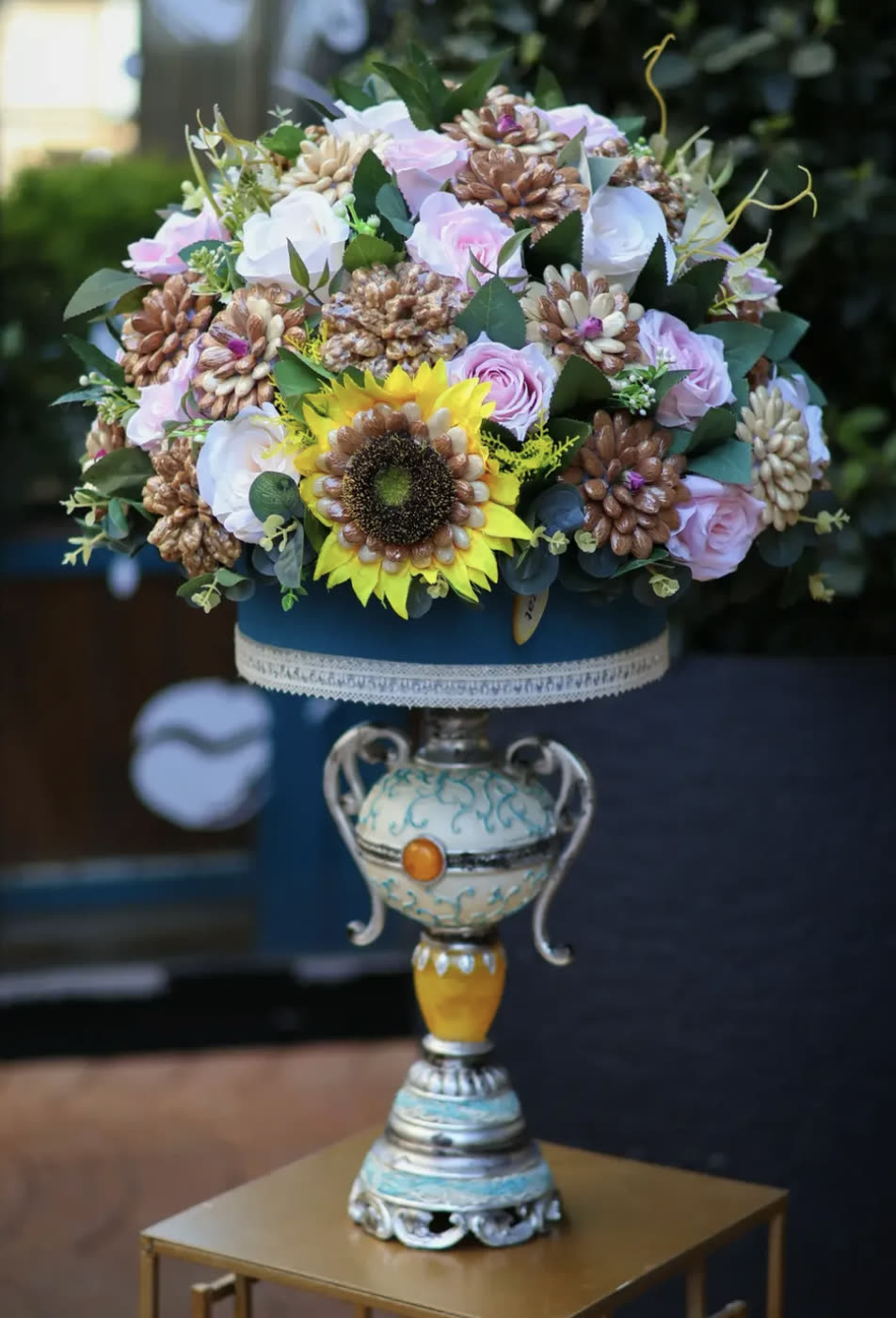 Tall urn of dried pine‑cone flowers, roses and a sunflower on a gold stand
